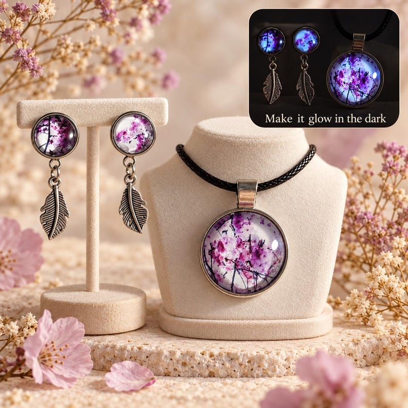 May include: A jewelry set featuring a pendant necklace and two drop earrings. Each piece has a circular glass cabochon with a purple and white floral print and a silver-toned metal feather charm. The necklace has a black cord. Text reads: Make it glow in the dark.