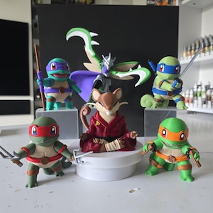 May include: A collection of five small plastic figurines featuring four turtle characters with coloured masks and weapons, and one central rodent character in a red robe sitting in a meditative pose. A winged figure stands behind them.