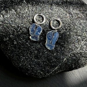 May include: A pair of silver hoop earrings featuring translucent, pale blue, irregular-shaped stone pendants. The earrings are composed of a circular metal hoop attached to a smooth, semi-transparent blue stone with a natural, organic, and uneven form.