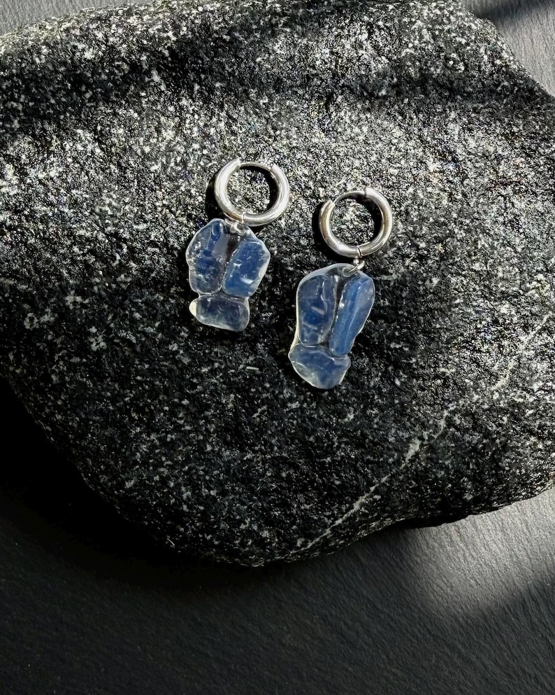 May include: A pair of silver hoop earrings featuring translucent, pale blue, irregular-shaped stone pendants. The earrings are composed of a circular metal hoop attached to a smooth, semi-transparent blue stone with a natural, organic, and uneven form.