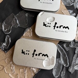 May include: Three rectangular silver metal tins with rounded corners, each featuring the black printed text "kii form by Arisha Kii" on the lid. The tins are arranged among scattered, irregular, translucent clear glass-like fragments.