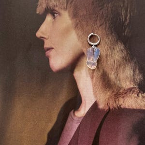 May include: A silver hoop earring featuring a dangling, irregular, translucent stone pendant with a pale blue and white iridescent sheen. The earring is displayed against a printed image of a person in profile, highlighting the stone's reflective surface.