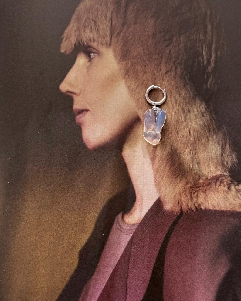 May include: A silver hoop earring featuring a dangling, irregular, translucent stone pendant with a pale blue and white iridescent sheen. The earring is displayed against a printed image of a person in profile, highlighting the stone's reflective surface.
