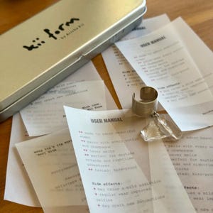 May include: A silver-toned metal cuff ring featuring a clear, irregular-shaped crystal accent. The ring is displayed alongside several white paper sheets labeled USER MANUAL and a rectangular silver metal storage tin marked kii form by Arisha Kii.