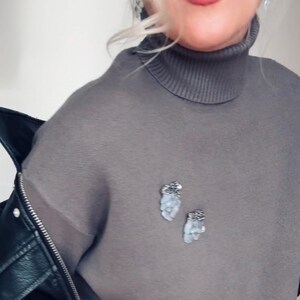May include: Two small, irregular-shaped brooches pinned to a grey turtleneck sweater. Each brooch features a textured, dark metallic top section and a cluster of pale, translucent, light blue stones or beads hanging below in a vertical arrangement.