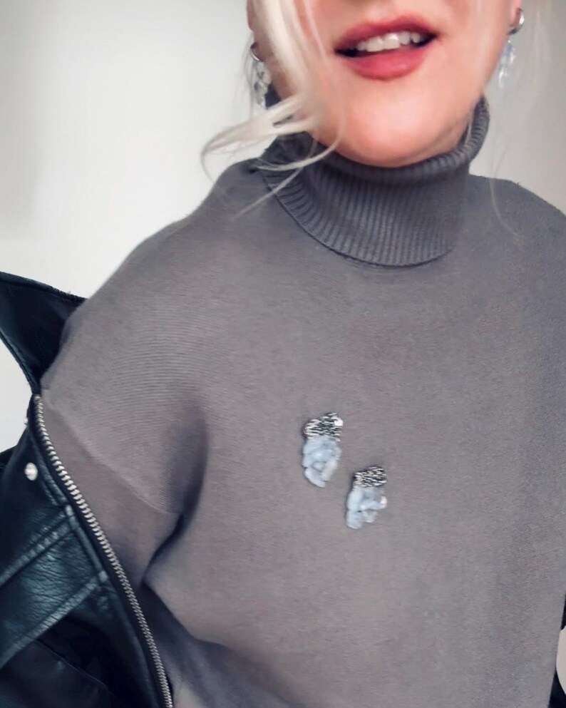 May include: Two small, irregular-shaped brooches pinned to a grey turtleneck sweater. Each brooch features a textured, dark metallic top section and a cluster of pale, translucent, light blue stones or beads hanging below in a vertical arrangement.
