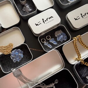 May include: A collection of silver-toned metal tins containing jewelry pieces. The tins feature the text kii form by Arisha Kii. Inside are various items including silver chains, hoop earrings, and translucent blue stone-like accents.