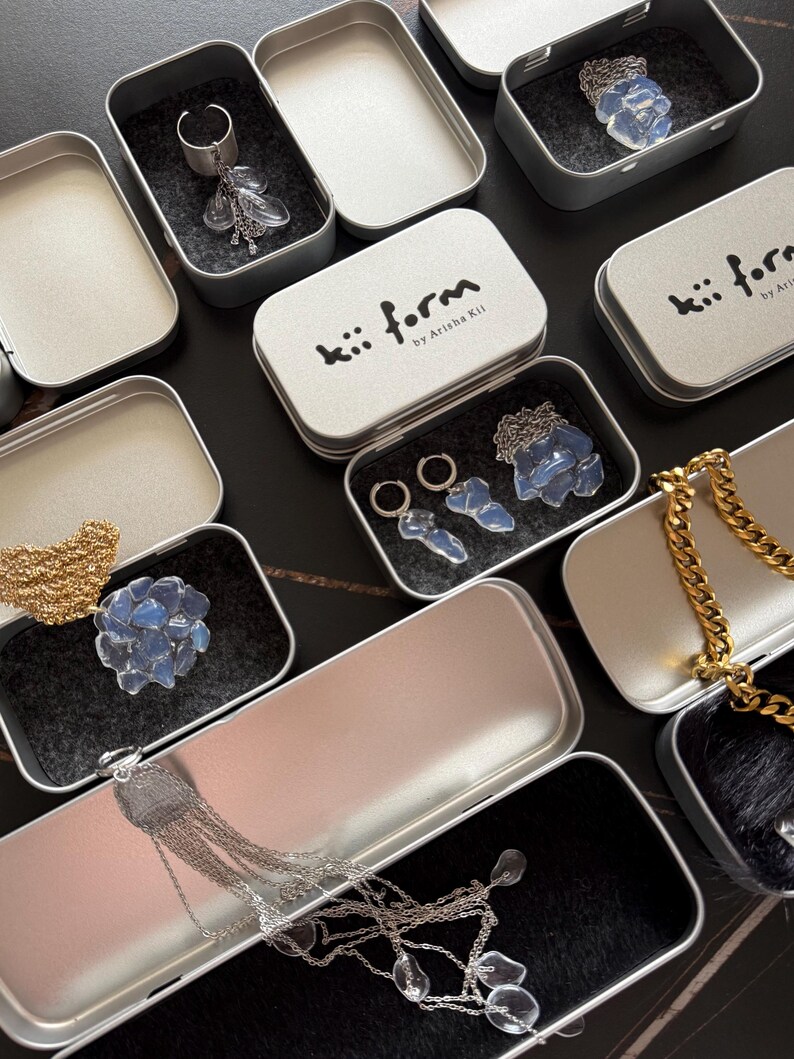 May include: A collection of silver-toned metal tins containing jewelry pieces. The tins feature the text kii form by Arisha Kii. Inside are various items including silver chains, hoop earrings, and translucent blue stone-like accents.