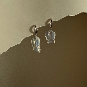 May include: A pair of silver hoop earrings featuring irregular, translucent, white-toned organic-shaped pendants that hang from the bottom of each hoop. The pendants have a smooth, glossy surface that reflects light with subtle blue and pearlescent hues.