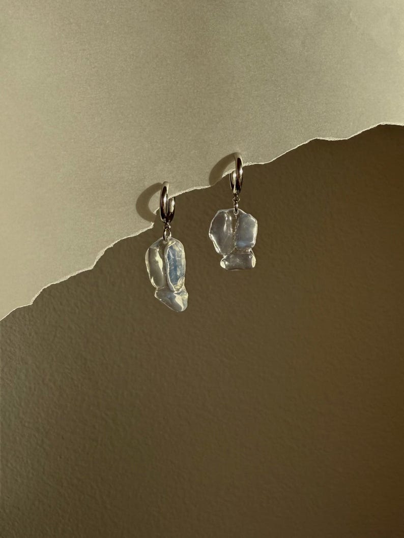 May include: A pair of silver hoop earrings featuring irregular, translucent, white-toned organic-shaped pendants that hang from the bottom of each hoop. The pendants have a smooth, glossy surface that reflects light with subtle blue and pearlescent hues.