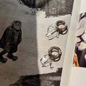 Puede incluir: Pendientes plateados con un dije transparente y texturizado en forma de hoja. Los pendientes tienen un diseño circular y se exhiben en una página de libro vintage con una ilustración en blanco y negro.