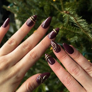 May include: Close-up of manicured nails painted a deep burgundy color. Some nails feature gold-colored embellishments, including rings and decorative accents. The nails are almond-shaped, and the background is a blurred Christmas tree.