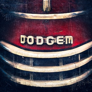 May include: Close-up of a vintage truck grille with the word "DODGEM" in silver letters on a red background. The grille has horizontal chrome bars and a weathered, textured appearance, suggesting an antique vehicle.