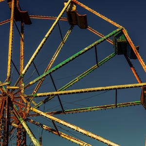 May include: An aged Ferris wheel against a deep blue sky. The metal structure shows signs of rust and wear, with faded green and yellow paint. The seats are visible, adding to the vintage amusement park theme.