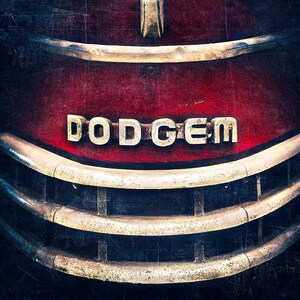 May include: Close-up of a vintage truck grille with the word "DODGEM" in silver letters on a red background. The grille has horizontal chrome bars and a weathered, textured appearance, suggesting an antique vehicle.