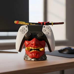 Op de afbeelding: Een wit-zwarte gamecontroller rust op een rood-gouden Oni-maskerstandaard, met een decoratief katana-zwaard erbovenop. De controller heeft witte zijpanelen en zwarte knoppen. Het masker heeft gouden tanden en een gedetailleerd ontwerp.