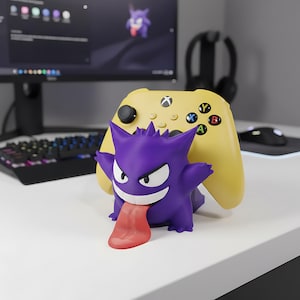 May include: A purple Gengar figure with a red tongue stands in front of a yellow Xbox controller. The controller has black buttons with coloured letters. The figure is on a white surface, with a computer and keyboard in the background.