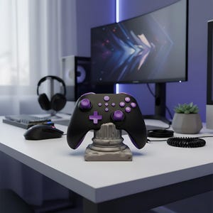 Può includere: Un controller di gioco nero e viola con un pulsante a forma di croce e pulsanti circolari, appoggiato su un supporto decorativo grigio. Il controller è su una scrivania bianca con un monitor del computer, cuffie e un mouse sullo sfondo.