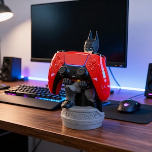 Könnte beinhalten: Ein Batman-Controller-Halter. Der Halter ist eine Statue von Batman, der einen rot-schwarzen Gamecontroller hält. Der Sockel ist grau mit einem dekorativen Design. Der Controller hat eine Standardgröße.