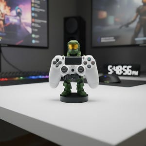 以下が含まれることがあります： 白いPlayStation 4コントローラーを持つ、緑と金のマスターチーフのフィギュア。フィギュアは黒い台座にあり、コントローラーは中央に配置されています。背景にはコンピューターモニターとキーボードが含まれています。