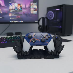 Op de afbeelding: Een blauw-witte gamecontroller met oranje accenten, rustend op een zwarte kristalvormige standaard. De controller heeft een kruisvormig D-pad en meerdere knoppen. Een computermonitor, toetsenbord en koptelefoon zijn op de achtergrond te zien.