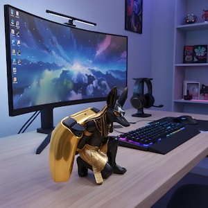 May include: A black and gold Anubis statue sits on a wooden desk in front of a curved computer monitor. The statue has a gold cape and accents. A keyboard, mouse, and headset are also on the desk.