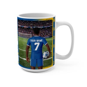 Peut inclure: Mug en c&eacute;ramique blanche avec un joueur de football en uniforme bleu portant le num&eacute;ro 7 et le texte "YOUR NAME". L'arri&egrave;re-plan montre un stade rempli de spectateurs. Le mug a une bordure bleue et jaune avec le mot "MADRID".