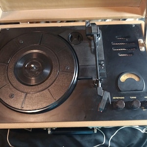 May include: A vintage portable record player in a tan and black case. The turntable has a black platter and tone arm. Controls include volume, tuning, and AM/FM/Phono. The case is open, revealing the player's components.