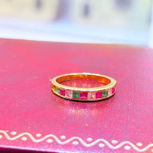May include: A gold ring with a band of alternating red, green, and clear square-cut gemstones. The ring is set against a red background.