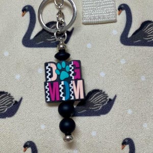 May include: A silver keychain with a charm that reads "DOG MOM" in pink, teal, and black. The charm features a paw print and is attached to a chain with black beads. The background is a fabric with swan patterns.