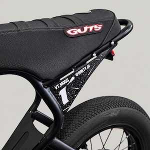 May include: A black motorcycle seat with a red and white GUTS logo patch. Below the seat, a black side panel features a white number 1, the text VT.MODS, and @BRETY.01 against a black background with white speckles resembling stars.