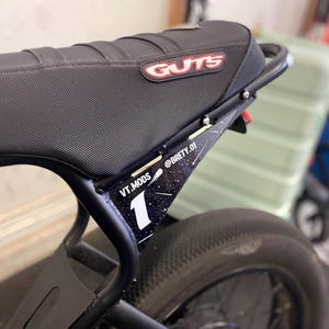 May include: A black motorcycle seat with a textured surface and a red and white GUTS logo. Below the seat, a black side panel features a white number 1, a comet graphic, and the text VT.MODS and @BRETY.01 against a black starry background.