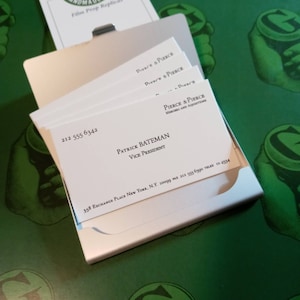 May include: A silver business card holder with several white business cards. The cards display the name "Patrick Bateman, Vice President" and the company "Pierce & Pierce". The background is a green surface with a repeating graphic.