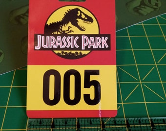May include: A rectangular Jurassic Park tag with a red top section and a yellow bottom section. The top section features the Jurassic Park logo with a dinosaur silhouette. The number "005" is printed in black on the yellow section.