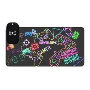 May include: A black gaming mouse pad with a wireless charging pad. The pad features neon-style illustrations of game controllers, a target, and headphones. Text includes "Gamer", "Level Up", and "Game Over".