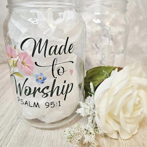 Puede incluir: Dos tarros de cristal transparentes con las palabras "Made to Worship" y "Salmo 95:1" en escritura negra, decorados con detalles florales rosas y azules. Una rosa blanca y una ramita de flores blancas están en primer plano.