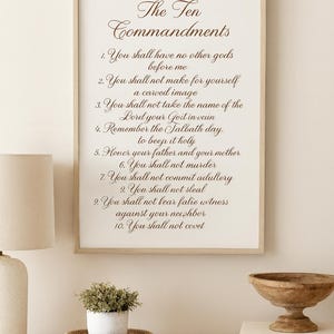 May include: A framed print displaying "The Ten Commandments" in elegant script. The text is centered on a cream-colored background within a light wood frame. The image is displayed on a light-colored wall, with a lamp, plant, and decorative bowl.