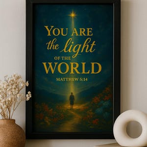 May include: Framed art print with the text "YOU ARE the light OF THE WORLD" and "MATTHEW 5:14". The image depicts a person walking towards a bright light. The artwork is in a black frame and is displayed on a wooden surface.