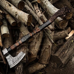 Könnte beinhalten: Eine handgefertigte Axt mit Holzgriff und Metallklinge. Der Griff ist mit schwarzem Leder umwickelt und weist aufwendige Schnitzereien auf. Die Klinge hat ein dekoratives Design und den Text "SCOCHED WOLF MENTALITY PREVAILS".