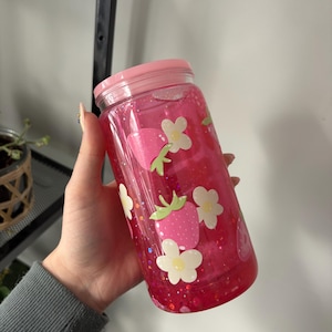 Peut inclure: Un gobelet rose transparent avec un couvercle rose, rempli de liquide rose et de paillettes. Le gobelet est décoré d'autocollants de fraises et de fleurs. Le gobelet est tenu par une personne.