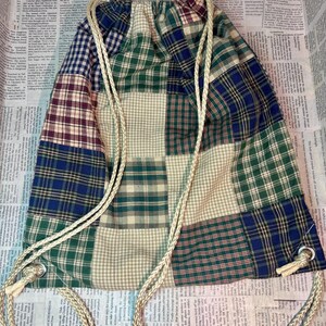 May include: A patchwork drawstring backpack featuring a variety of plaid patterns in shades of green, blue, and burgundy. The bag has a cream-colored drawstring closure and eyelets. The bag is made of fabric.