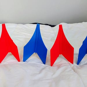 May include: Four abstract, geometric shapes in red and blue are arranged on a white surface. The shapes have a symmetrical design with a pointed top and a wide base. The color scheme is a vibrant contrast.