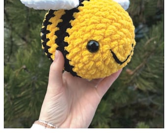 Puede incluir: Una abeja de ganchillo hecha a mano con detalles amarillos, negros y blancos. La abeja tiene una cara sonriente y está sostenida en una mano. El texto "Crochet Bee Pattern" se muestra debajo.