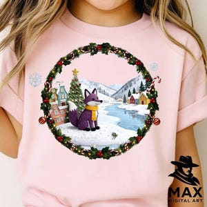 May include: A light pink t-shirt featuring a winter scene with a purple fox wearing a yellow scarf. The fox is inside a wreath with holly and red berries. A Christmas tree, houses, and a snowy landscape are in the background.