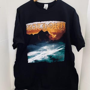 Op de afbeelding: Zwart T-shirt met een grafisch ontwerp met het woord "Bathory" in een oranje en gouden lettertype boven een berglandschap met een stormachtige oceaan. De woorden "Twilight of the Gods" staan eronder.