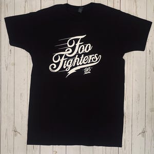 Może przedstawiać: Czarny t-shirt z białym napisem "Foo Fighters" w stylizowanej czcionce, błyskawicą i rokiem "95". Koszulka wykonana jest z miękkiego materiału i ma krótkie rękawy.