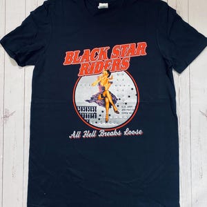 Op de afbeelding: Zwart T-shirt met de tekst "BLACK STAR RIDERS" in oranje en wit. Het shirt heeft een afbeelding van een vrouw in een paarse jurk in een cirkel. De tekst "All Hell Breaks Loose" staat eronder.