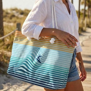 Pode incluir: Uma bolsa de praia listrada com um gráfico de surfista. A bolsa apresenta listras horizontais em tons de azul, verde e laranja. Possui alças de corda branca e está sendo carregada ao ar livre.