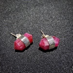 Może przedstawiać: Para kolczyków sztyftów z fasetowanymi, głęboko czerwonymi kamieniami szlachetnymi. Każdy kamień jest utrzymywany na miejscu przez srebrną opaskę. Kolczyki są prezentowane na teksturowanej, ciemnoszarej powierzchni.