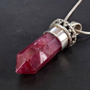 May include: A faceted, deep red crystal pendant with a silver-tone metal setting and bail. The pendant hangs from a delicate silver chain. The crystal is a rich, saturated colour, and the metal has an ornate design.